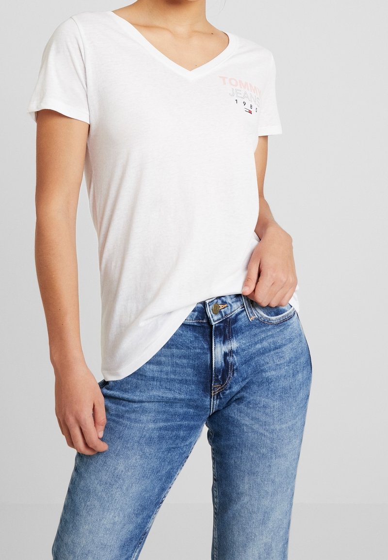 Camiseta de algodón blanca con escote en V y mangas cortas. Presenta "TOMMY JEANS" impreso en rosa. Combinada con jeans de mezclilla azul.