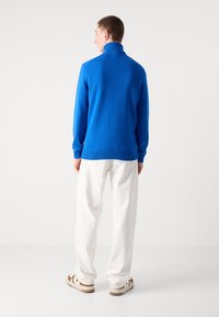 Blauer Strickpullover mit hohem Kragen, kombiniert mit weißen Hosen und lässigen Schuhen, mit einem lockeren Schnitt und gerippten Bündchen.