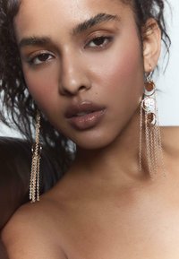 Zlaté náušnice s kruhovými kovovými akcentmi a reťazami, s lesklým povrchom. Modelka má prirodzený make-up a kučeravé vlasy.