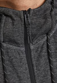 Sudadera con cremallera en tejido gris oscuro con un acabado texturizado. Presenta un cuello alto, cordones ajustables y acentos de costura blanca en contraste.