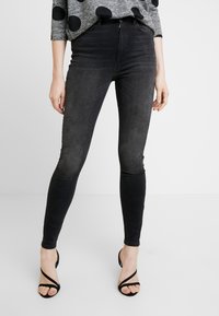 Jeans negros ajustados exhibidos en un modelo, con una cintura alta, textura suave y diseño elegante, combinados con tacones negros con tiras.