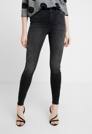 Jeans negros ajustados exhibidos en un modelo, con una cintura alta, textura suave y diseño elegante, combinados con tacones negros con tiras.