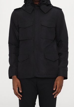 Homme portant une veste noire à capuche avec quatre poches avant à rabat et un pantalon noir, debout devant un fond uni.