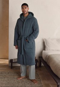 Gepolsterter dunkelblauer Morgenrock mit Kapuze, Bindegürtel und zwei Fronttaschen; kombiniert mit hell gestreiften Hosen. Raumgestaltung in neutralen Tönen.