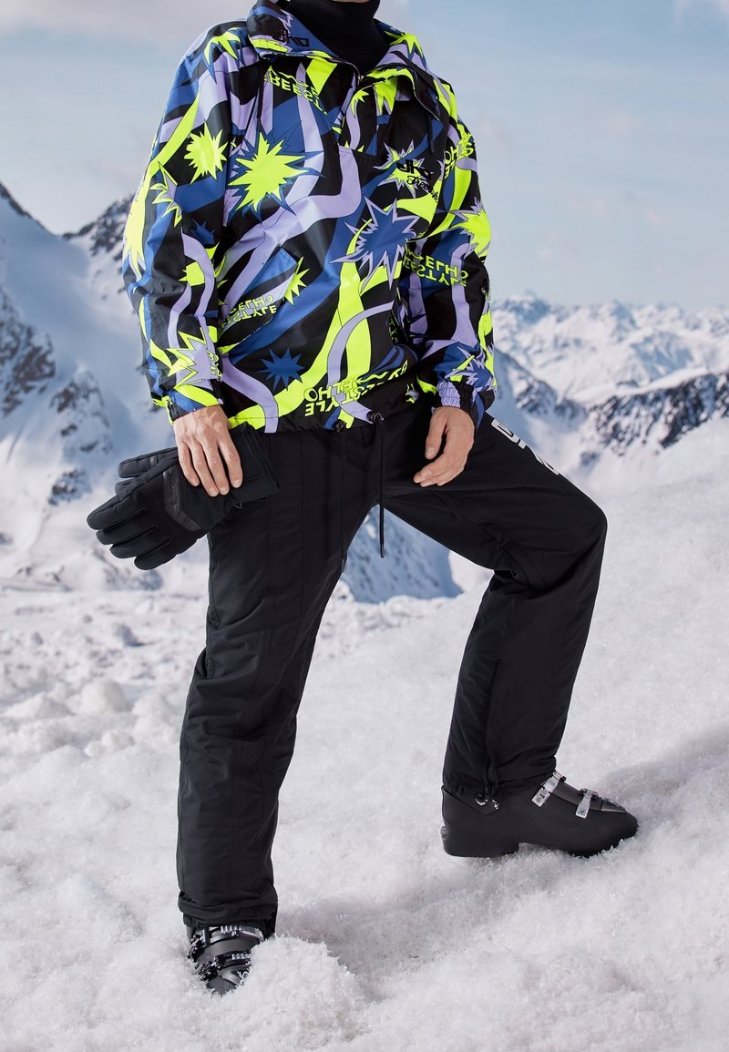Personne portant une veste colorée à motifs, un pantalon de neige noir et des chaussures de ski, debout sur une montagne enneigée avec des gants à la main.