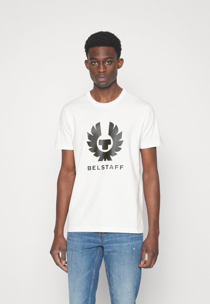 Balta kokvilnas t-kreklu ar melnu logo ar spārniem un tekstu "BELSTAFF" zem tā, ar apaļu kaklu, īsām piedurknēm un gaiši ziliem džinsiem.