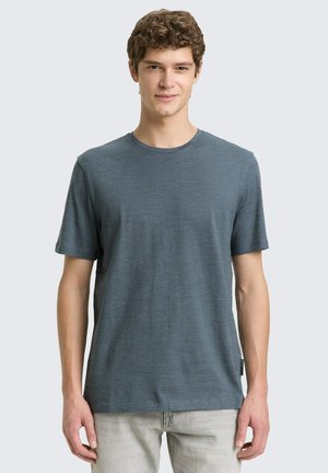 Młody mężczyzna z kręconymi włosami, ubrany w gładki niebieski t-shirt z krótkim rękawem i jasne spodnie, stojący na jednolitym tle.