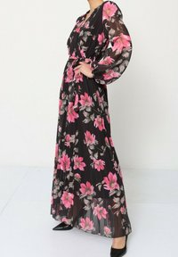 Robe longue noire à motifs floraux roses et gris, avec des manches longues et transparentes et une taille nouée, assortie à des chaussures noires.
