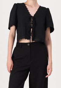 Svart kort topp i mjukt tyg med en scalloped V-hals, korta ärmar och en knytning framtill med dekorativa kanter.