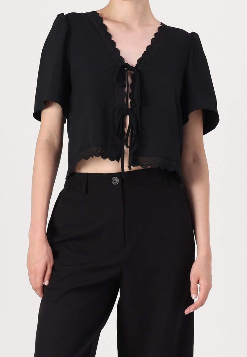Svart kort topp i mjukt tyg med en scalloped V-hals, korta ärmar och en knytning framtill med dekorativa kanter.