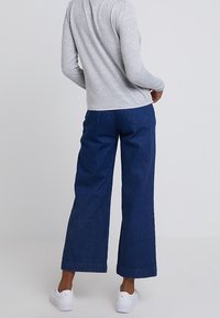 Pantalones vaqueros de pierna ancha en azul oscuro, tela de algodón con dobladillo sin rematar. Combinados con una camiseta de manga larga gris y zapatillas blancas.