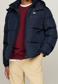 Mörkblå puffjacka med dragkedja och huva, prydd med en logotyp på bröstet och ribbade muddar, lagd över en vinröd sweatshirt med rund hals.