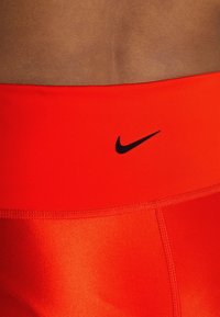Červené sportovní šortky s hladkou texturou, se známým černým logem Nike na pase a jemnými detaily šití.
