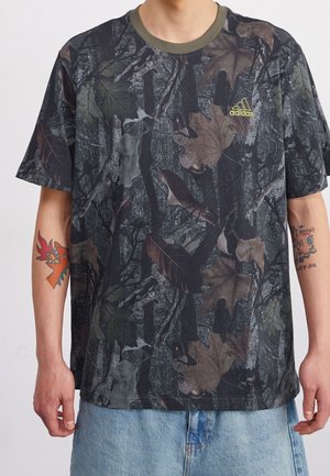 Mężczyzna noszący krótką, moro-patterned koszulkę Adidas z zielonym kołnierzem i dżinsy, z widocznymi tatuażami na obu rękach.