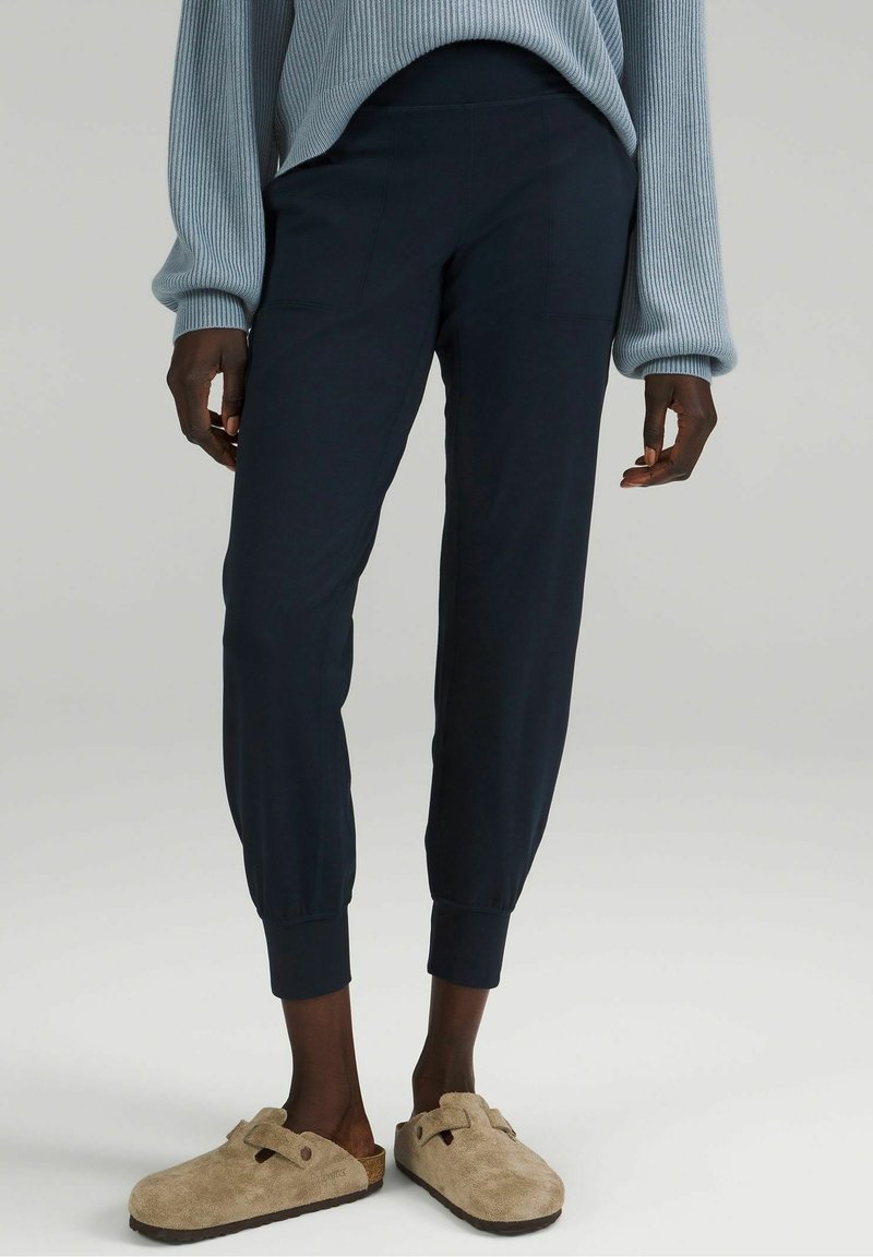 Pantalons de jogger de couleur marine avec des poignets élastiques et des poches latérales, fabriqués à partir d'un matériau doux. Associés à des chaussures beiges à enfiler.