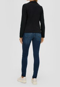 Musta pitkähihainen takki, jossa on kaulus, yhdistettynä tummansinisiin slim fit -farkkuihin ja valkoisiin lenkkareihin. Takissa on sileä pinta ja räätälöity istuvuus.