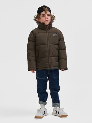 Kind draagt een bruine donsjas, opgerolde blauwe spijkerbroek, witte sneakers en een zwarte pet achterstevoren, staand tegen een effen achtergrond.