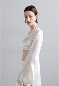 Costum din satin alb format din două piese, cu un top scurt cu mâneci lungi, transparente și pantaloni cu talie înaltă, textură moale și design fluid.