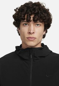 Zwarte jas met rits, uitgevoerd met een capuchon met trekkoord en lichtgewicht stof. Een klein zwart Nike-logo is geplaatst op de linkerborst.