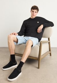 Zwarte sweatshirt met een logopatch, lichtblauwe denim shorts, zwarte sneakers met witte zolen en geribde zwarte sokken, zittend op een beige stoel.