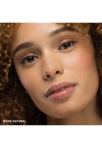 Produit de maquillage étiqueté "Naturel Chaud" appliqué sur le visage, avec des cheveux bouclés, une peau lisse, des teintes pêche douces sur les paupières et des lèvres brillantes.