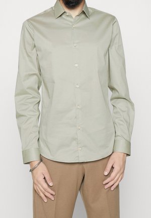 Homme portant une chemise vert clair à boutons et un pantalon marron, debout les mains le long du corps devant un fond uni.
