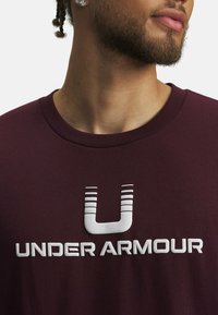 Mørkerød T-shirt med korte ærmer lavet af bomuldsblanding, med et stort hvidt "UA" logo og teksten "UNDER ARMOUR" foran. Glat tekstur.