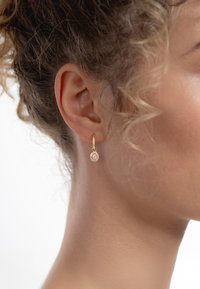 Boucle d'oreille en or en forme de créole avec un petit pendentif rond orné de pierres claires, attachée à une oreille texturée. Le fond est blanc uni.