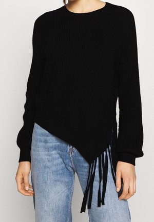 Donna che indossa un maglione nero lavorato a maglia con orlo asimmetrico e dettagli con frange abbinato a jeans azzurri chiari.