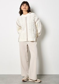 Biały sweter z dzianiny z wzorem ażurowym i beżowymi akcentami, w połączeniu z jasnobeżowymi spodniami szerokimi w nogawkach i beżowymi butami wsuwanymi.