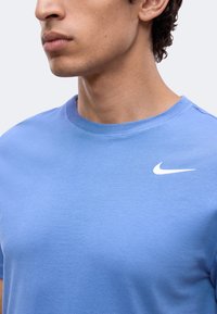 Homme portant un t-shirt Nike bleu clair à col ras-du-cou avec un logo Nike blanc sur la poitrine gauche, visible des épaules à la bouche.
