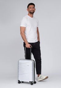 Silberner Hartschalenkoffer mit strukturiertem, diagonalem Linienmuster, ausziehbarem Griff und vier Rädern. Benutzer hält den Koffer und trägt legere Kleidung.