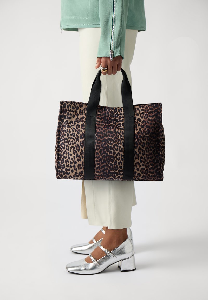 Tote-väska i leopardmönster med svarta handtag, tillverkad av texturerat tyg, har en rymlig design och robusta sömmar. Bärs av en person.