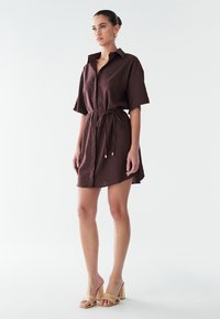 Brun skjortekjole med knapper, bælte i taljen, korte ærmer og en let buet kant forneden. Båret med beige hælede sandaler.