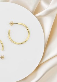 Guldtonade hoopörhängen med en tvinnad design, med en säker bakre stängning. Visade på en rund vit yta mot en satinbakgrund.