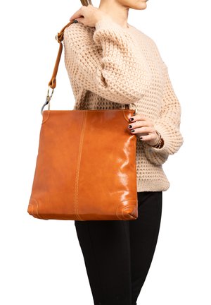 Vrouw in beige trui die een grote lichtbruine lederen schoudertas vasthoudt met metalen ringen en verstelbare riem.