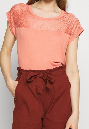 Kobieta w koralowym topie z krótkim rękawem i koronką na karczku oraz wysokim stanem spodni w kolorze rdzy, zawiązanych na materiałowy pasek.