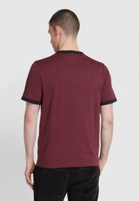 Uomo visto di spalle che indossa una t-shirt cremisi con rifiniture nere sul colletto e sulle maniche, e pantaloni neri, su uno sfondo semplice.