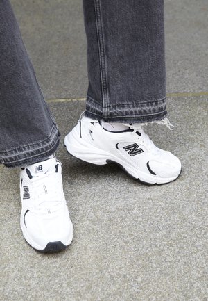 Białe sneakersy New Balance mają cholewkę z siateczki i syntetyku z czarnymi akcentami. Noszone z szarymi dżinsami z postrzępionym dołem na teksturowanej powierzchni.