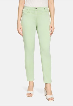 Frau trägt hellgrüne, schmal geschnittene Hose mit Vordertaschen, kombiniert mit weißen, offenen Blockabsatz-Sandalen, steht vor weißem Hintergrund.