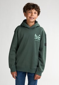 Groene hoodie met een voorzak, capuchon met trekkoord en een logo met bomen en bergen. Gecombineerd met blauwe jeans. Gemaakt van een zachte katoenmix.
