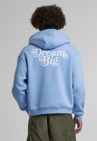 Svetlomodrá mikina s kapucňou a veľkým bielym nápisom "Dream Big" na chrbte. Vyrobená z mäkkého materiálu, s kapucňou a rebrovanými manžetami, v ležérnom dizajne.