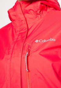 Veste Columbia rouge vif avec poche poitrine zippée et col haut, montrant la texture du tissu et les détails du logo.