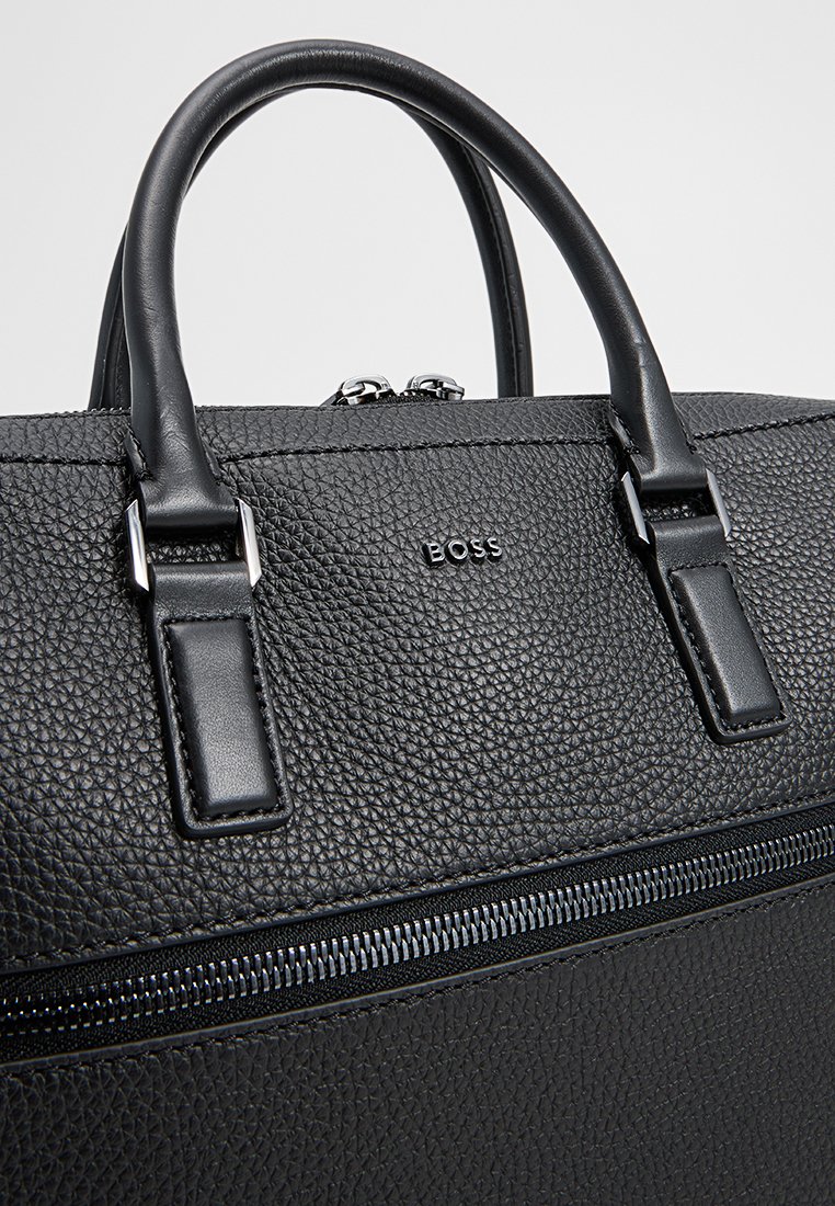 Malette en cuir noir avec une finition texturée, dotée de doubles anses supérieures, de quincaillerie argentée et d'une fermeture éclair centrale. Logo gravé à l'avant.