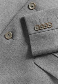 Šviesiai pilkas švarko audinys su rudos spalvos keturių skylių sagomis, turintis rankovę su keturiomis sagomis ir matoma priekyje esanti sagu.