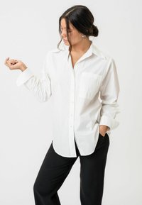 Fehér, könnyű szövetből készült ing, gallérral, egy zsebbel és hosszú ujjal, fekete nadrággal párosítva.