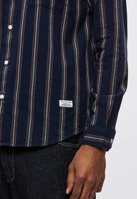 Granatowa koszula na guziki z pionowymi paskami w kolorach bordowym, białym i szarym. Posiada kieszeń na piersi oraz metkę marki przy dolnym brzegu.