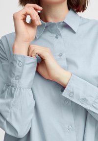 Light blue collared shirt with buttoned cuffs and gathered sleeve detailing. Smooth texture and a classic, tailored design.