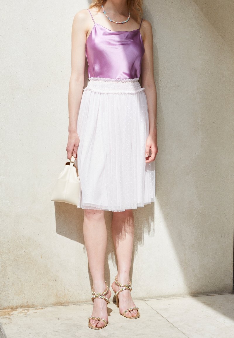 Camisole en satin lavande avec fines bretelles, associée à une jupe plissée blanche longueur genou avec des pois. Talons dorés à lanières et petit sac crème.