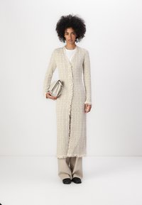 Un long cardigan beige clair en tissu tricoté texturé avec un ourlet à franges, doté d'une fermeture à boutons et de manches longues, porté avec un sac argenté.